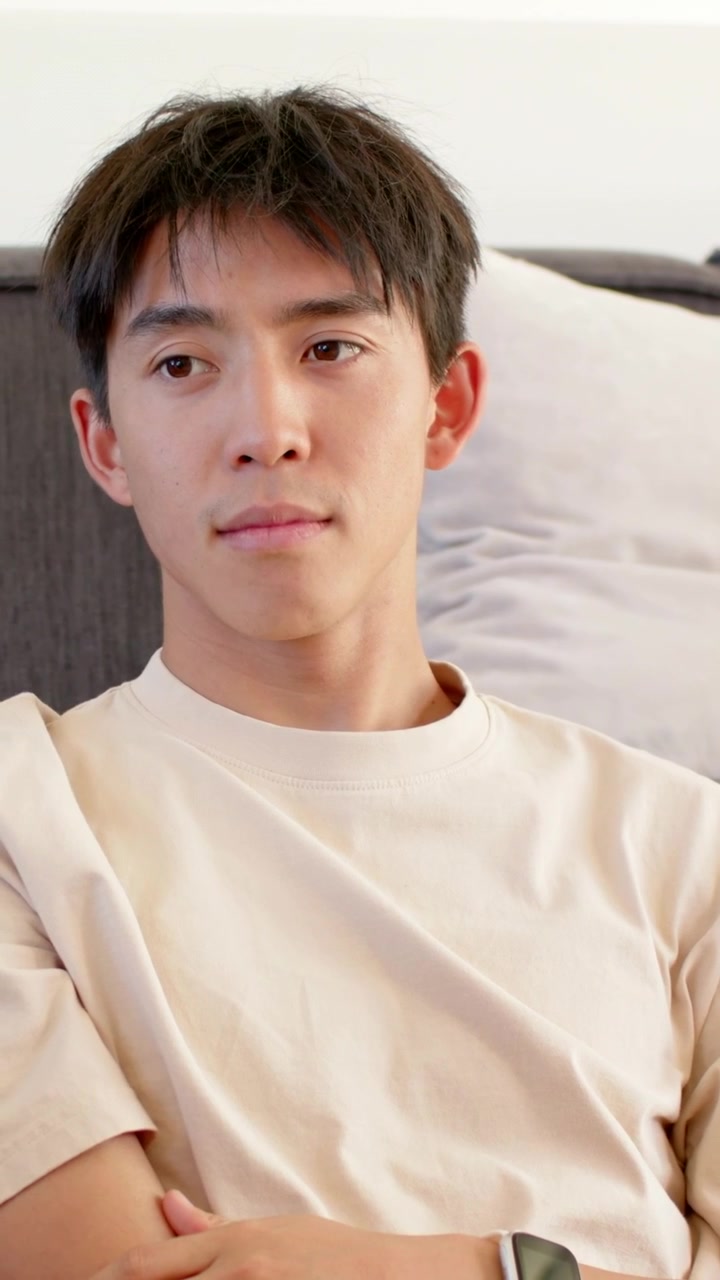 Video verticale: Si sente una voce fuori campo, un uomo asiatico con maglietta beige parla sul divano mostrando uno smartwatch.