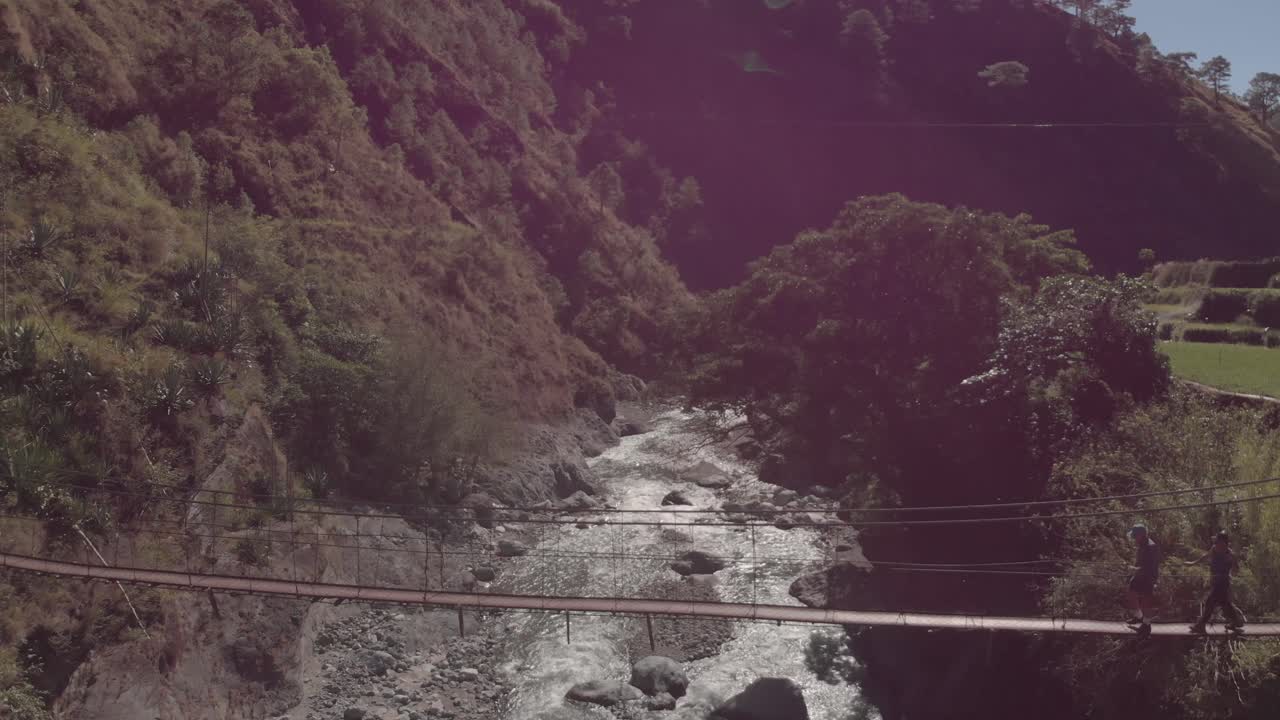 two men hiking walking across a metal wire suspension bridge spanning across a river in wild jungle mountain terrain surrounded by trees green light flair reversing aerial in benguet philippines