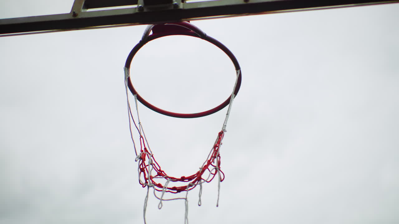 Canestro da basket e rete con gocce di pioggia che cadono dalla rete a catena, su uno sfondo di cielo grigio nuvoloso, un pallone sfiora il bordo e poi cade fuori dall'inquadratura, tabellone scolorito e vernice scrostata evocano l'atmosfera di un parco giochi urbano