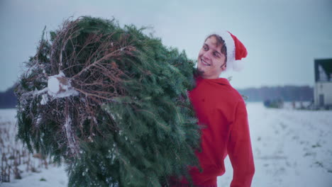 hombre feliz llevando el árbol de navidad mientras camina por el campo cubierto de nieve