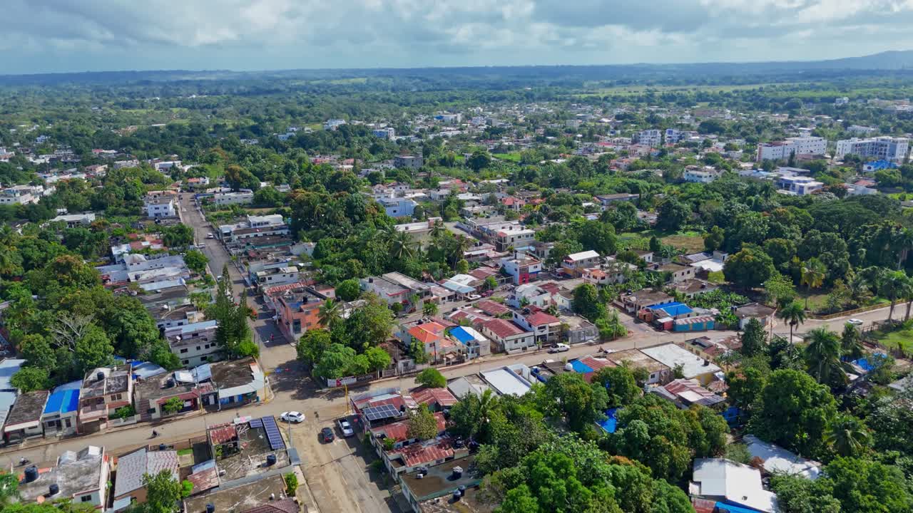 Aerial flyover Cotui Cityscape with Tropical palm trees and colored roofs at sunny day. Main street with traffic in town on Dominican Republic Island. Wide shot.