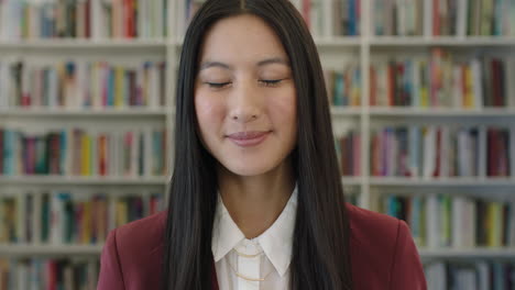 Retrato-De-Una-Linda-Joven-Estudiante-Asiática-Sonriendo-Confiada-Mirando-La-Cámara-En-El-Fondo-De-La-Estantería-De-La-Biblioteca-Pública