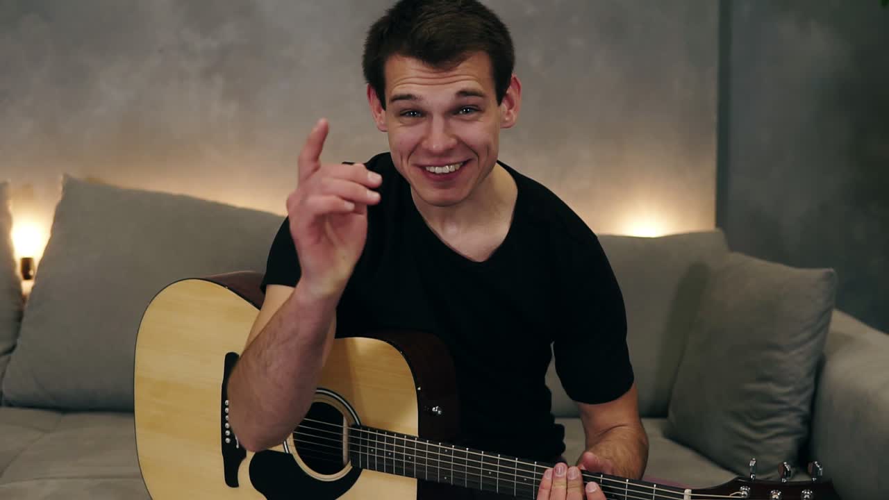 검은 티셔츠를 입은 짧은 머리카락의 백인 남자가 집에서 소파에 앉아 음악 악기에 대한 비디오 블로그를 만들거나 그의 추종자들을 위해 노래를 녹음하는 초상화. 카메라를 보고