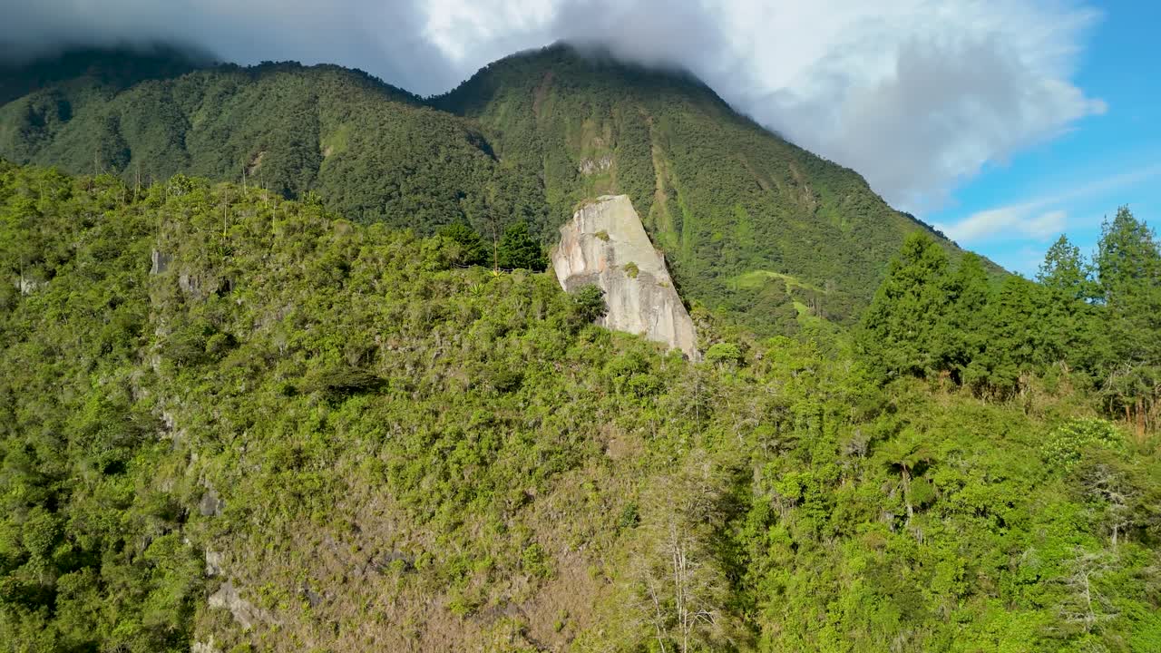 Plano aéreo en paralaje de la formación rocosa sagrada Piedra de la Luz, rodeada por un denso bosque verde en la ladera de una montaña bajo un cielo nublado en Baños, Ecuador.