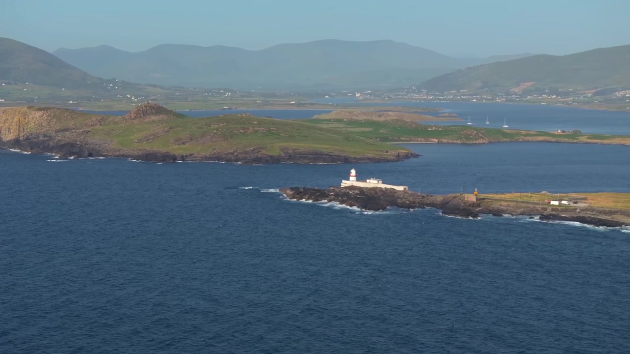 Il faro dell'isola di Valentia si trova su un promontorio basso mentre un drone scivola oltre un'ampia baia sostenuta da colline a strati e case sparse.