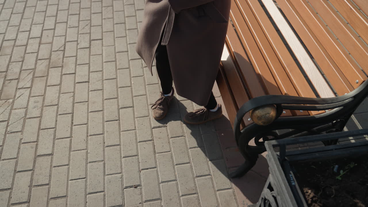 Kvinne i rød beret på en benk, reiseblogger prater i telefonen og smiler, i brun kåpe, sollys kaster lang skygge over en brosteinsbelagt parkscene, uformell samtale, ærlig hverdagsøyeblikk.