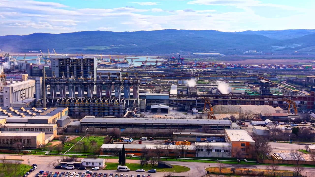 Bulgaristan'ın Varna şehrinde deniz kıyısına yakın bir sanayi tesisinin yüksekten görünümü. Büyük fabrika yapılarını, buhar emisyonlarını, hammaddeleri ve uzaktaki liman vinçlerini yakalayan perspektif.