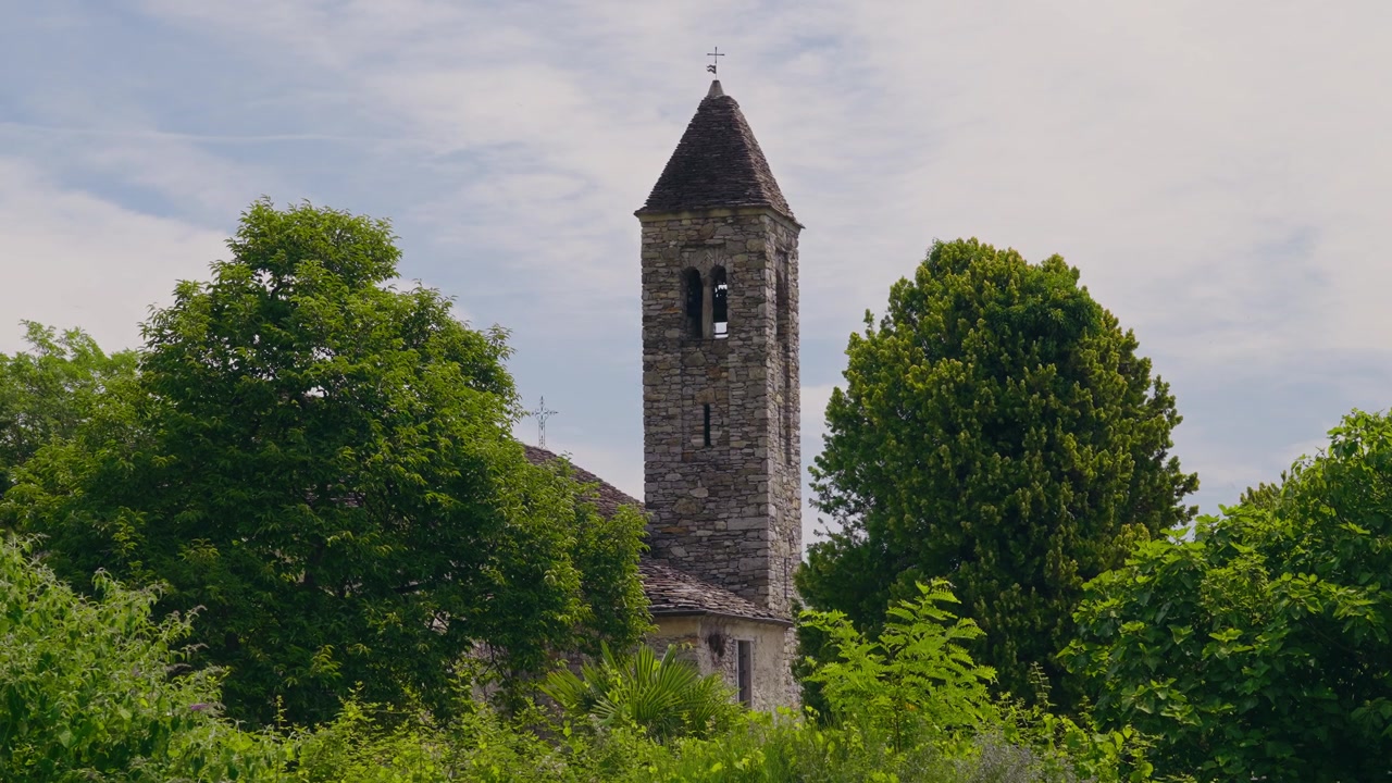 Malerischer steinerner Kirchturm, der über üppig grünen Bäumen aufragt, unter einem blauen Himmel mit sanften Wolken. Eine friedliche Sommerszene, die historische italienische Architektur und ländlichen Charme zeigt.