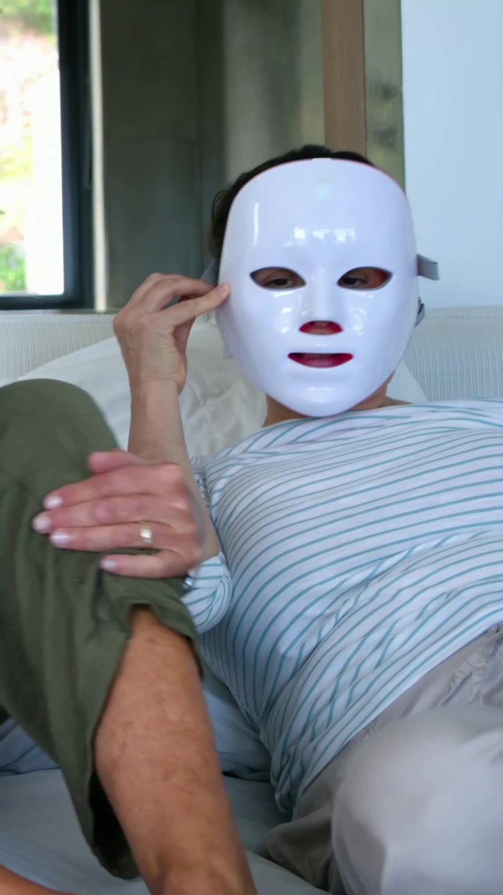 Vídeo vertical: Mulher de blusa listrada estendendo a mão e ajustando máscara de LED branca na cama após gesto de amiga.
