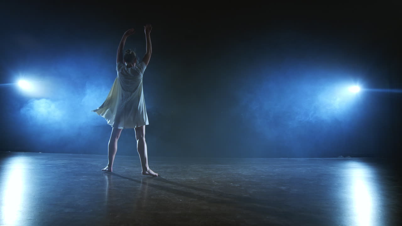 Dramatic spins and flips of a modern ballet dancer from a musical. A single woman emotionally dances on stage against a dark background with smoke in the spotlight in slow motion.