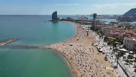 drone footage of barceloneta beach showing long curved shoreline, turquoise water and busy sandy coast. urban buildings and harbor visible in background under bright sunny sky