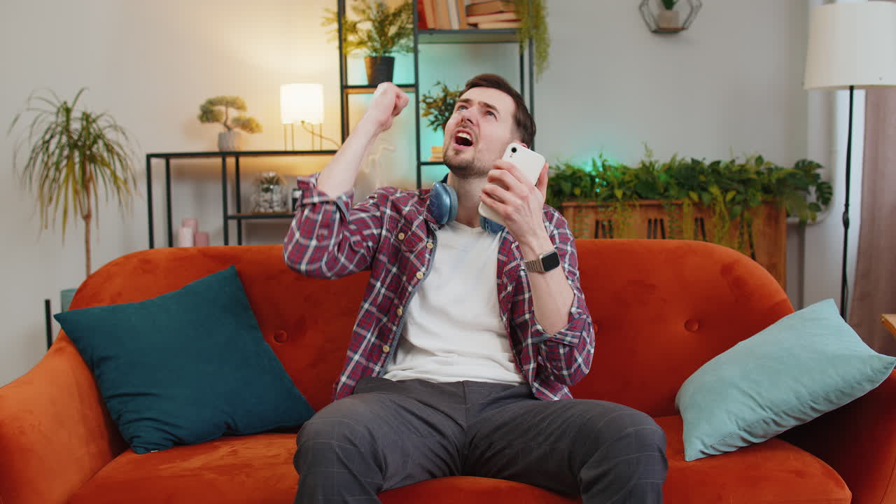 Irritated caucasian man with smartphone annoyed by noisy neighbors suffers from headache on sofa
