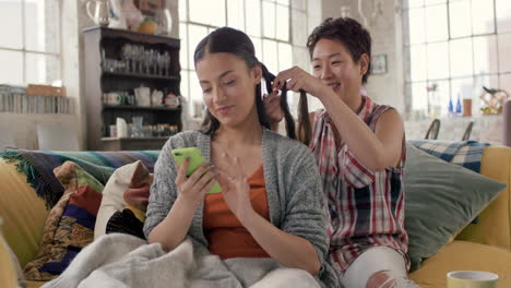 Multi-racial-girl-friends-braiding-hair-smiling-using-mobile-phone-hanging-out-on-sofa