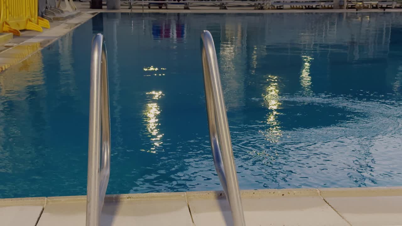 Still shot of quiet swimming pool with metal ladder and glowing water reflections at night