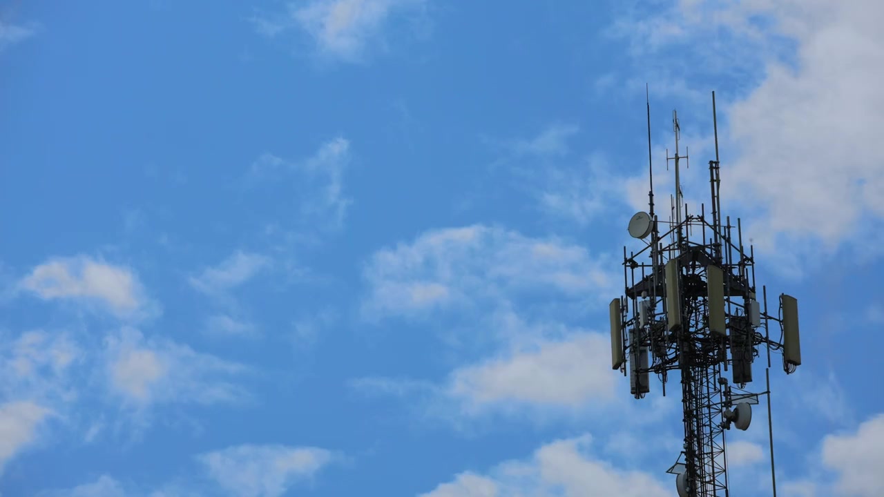 A timelapse video still showing a tall telecommunications cell tower with antennas. White clouds move dynamically across a bright blue sky in the background during daytime.