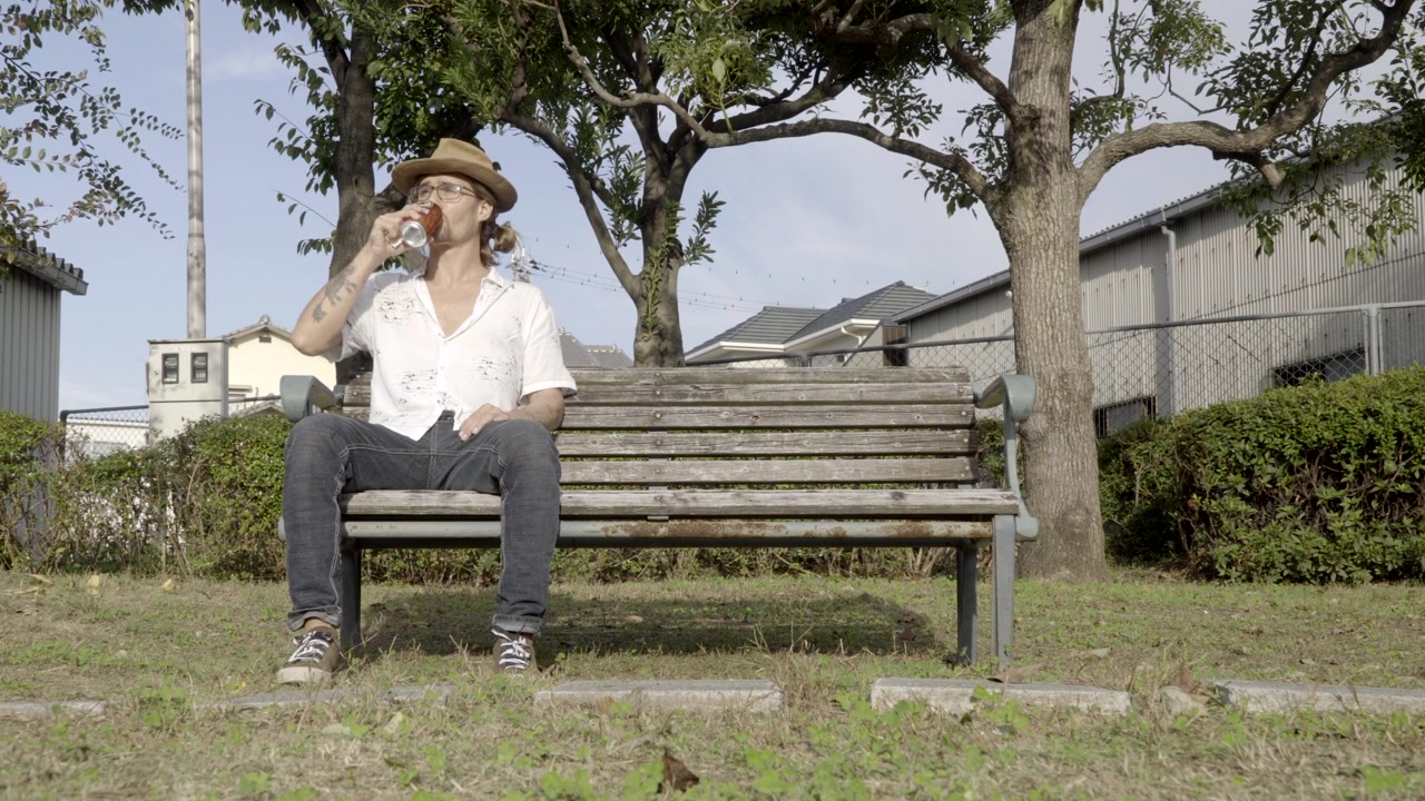 Un homme avec un chapeau et des lunettes, assis sur un banc dans le parc, se détend et sirote une bière.