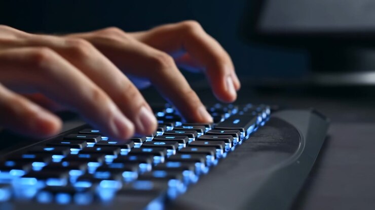 Nærbillede viser begge hænder, der bevæger sig over glødende tastaturtaster, med gentagne fingertryk, kølig blå belysning, lav fokus, mørk skrivebordsoverflade og sløret skærm bagved.