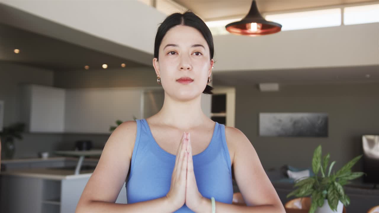 Donna asiatica in piedi a casa, con i palmi uniti, che respira, apre gli occhi e si concentra vicino a una lampada a sospensione.