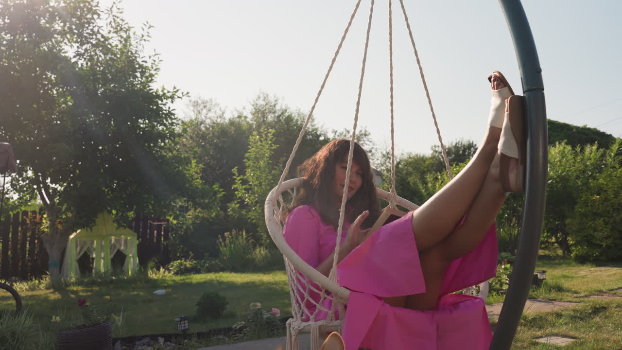 Garden Lounge Woman In Pink Swing With Dog At Feet On Wooden Deck, Relaxed Pose With Legs Up, Golden Light Filtering Through Trees, Cozy Pet Companionship And Summer Style Content