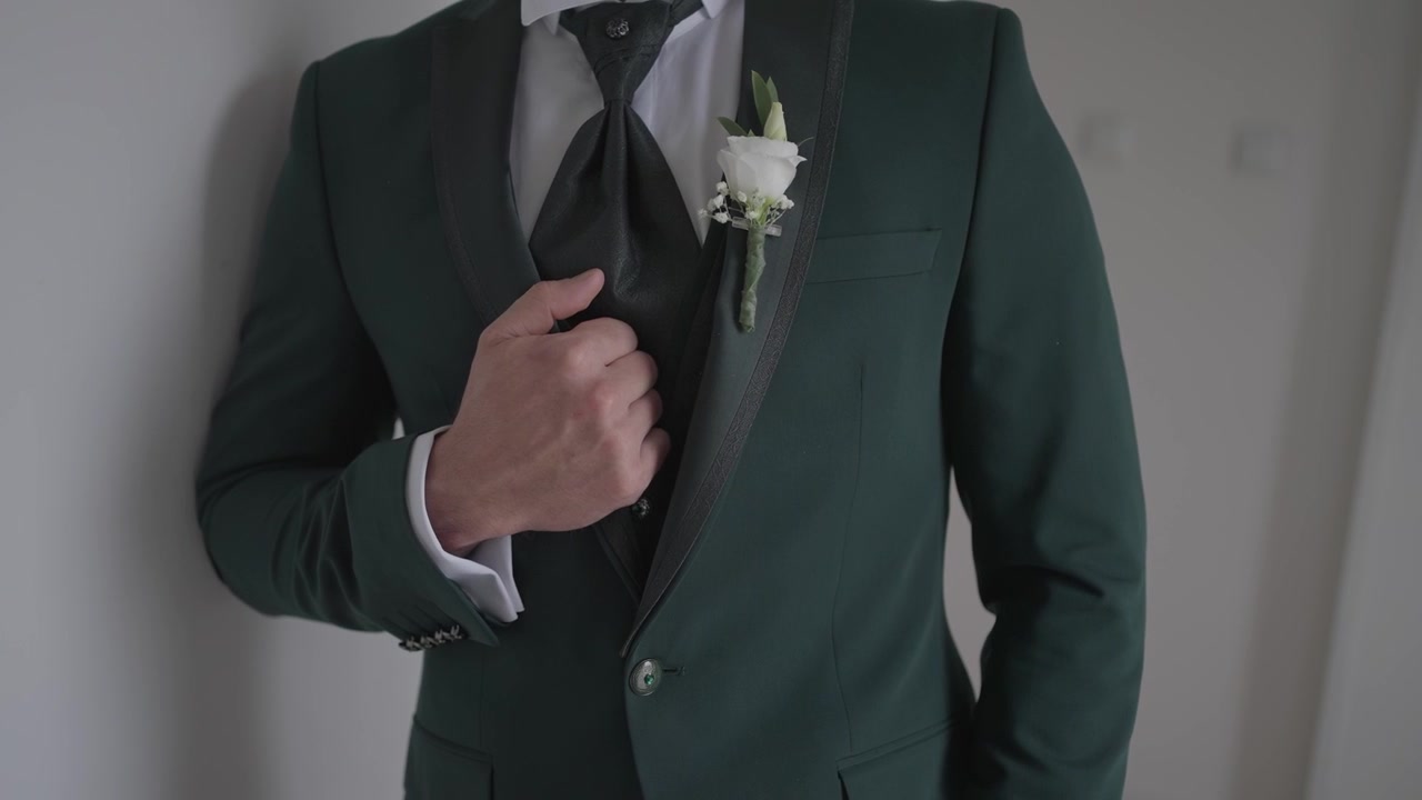 Primo piano della mano dello sposo che sistema cravatta e abito con la boutonnière al chiuso.