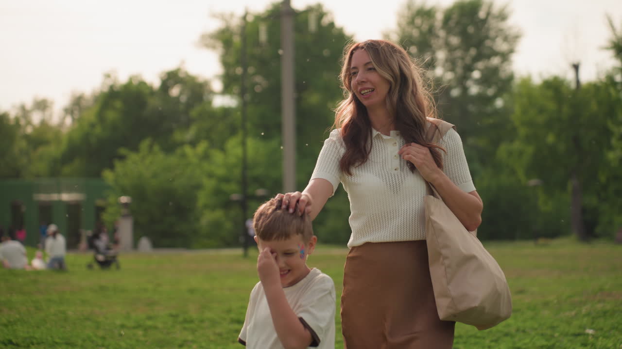 paint covered child holding mothers hand spinning round in park grassy field while mother watches smiling carrying tote bag hair blowing in breeze during sunny outdoor playful moment