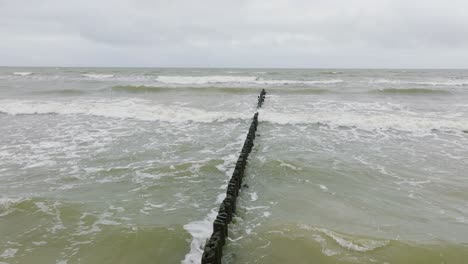 establishing aerial view of baltic sea coast on a overcast day, old wooden pier, white sand beach, large storm waves crushing against the coast, climate changes, wide drone shot moving forward low