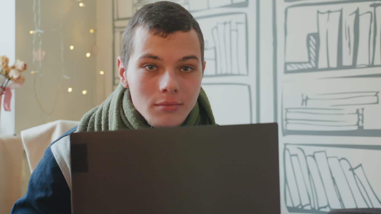 Caucasian man looking away from laptop thoughtful planner sketching ideas, scarf draped, fairy lights and bookshelf backdrop, moments of reflection, roles include strategist, designer, consultant