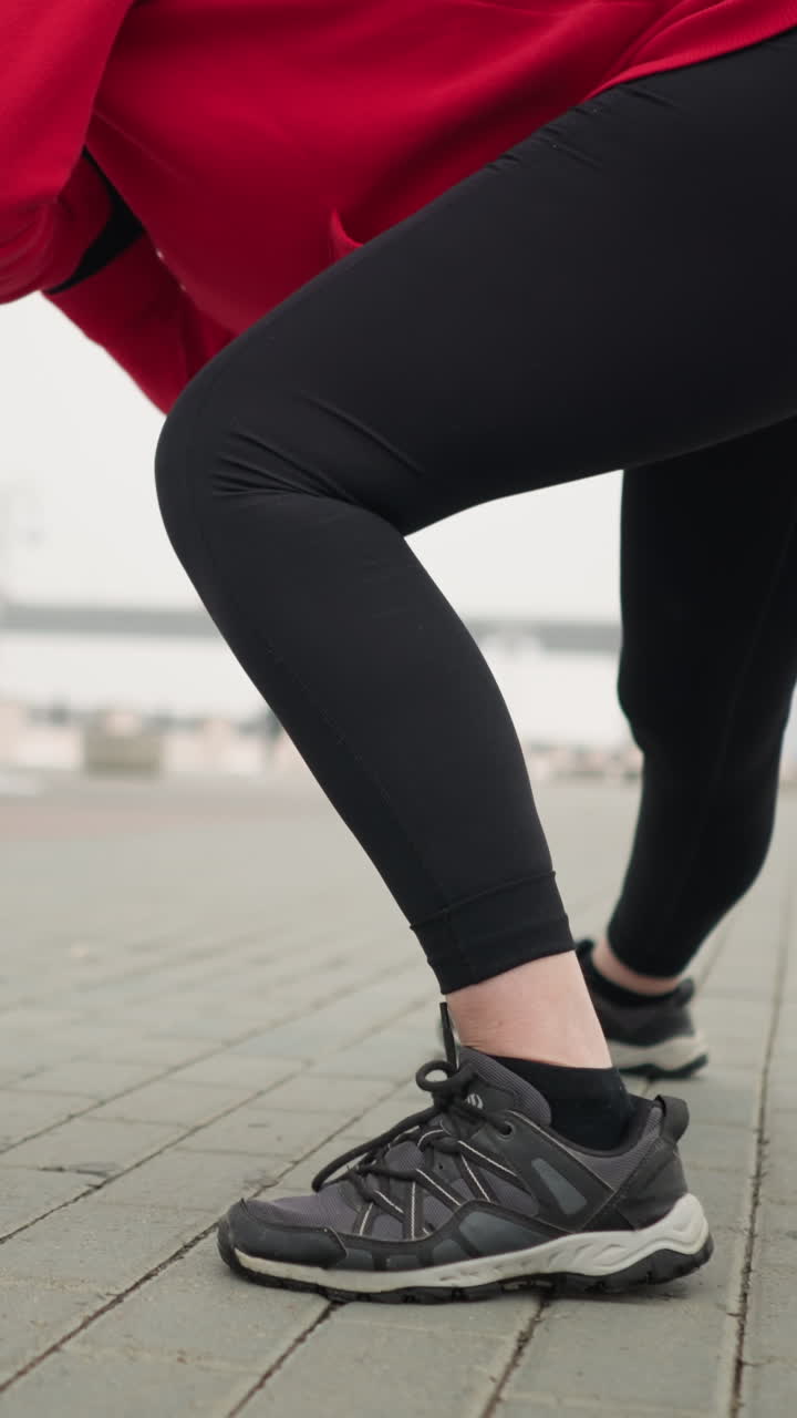Low-angle view of lady in leggings stretching into lunge during outdoor winter workout under modern canopy structure, with blurred pedestrian and distant bridge with cars in foggy urban surroundings