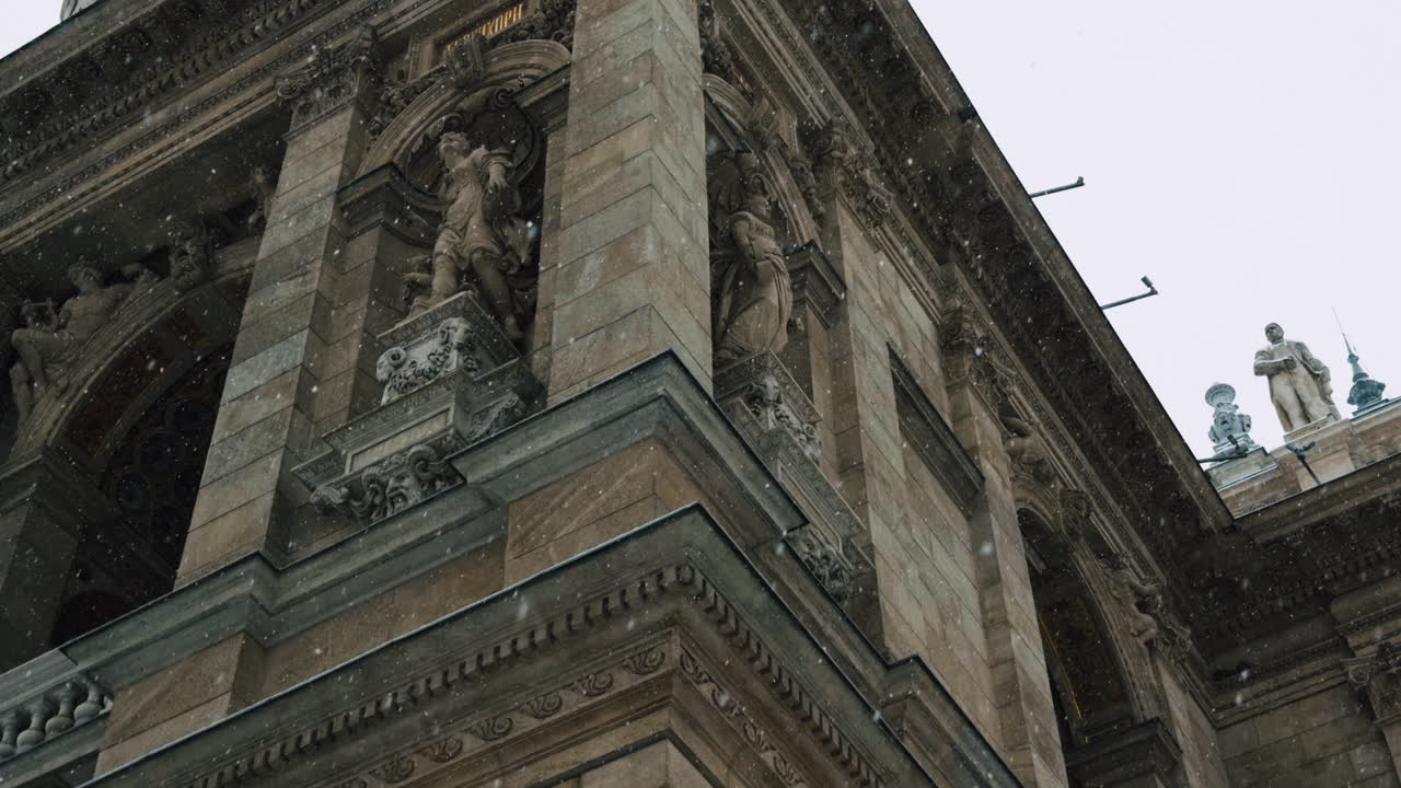Vista de perto da fachada da Ópera de Budapeste durante forte nevasca. Estátuas históricas, colunas de pedra e arquitetura clássica detalhada em uma atmosfera fria de inverno