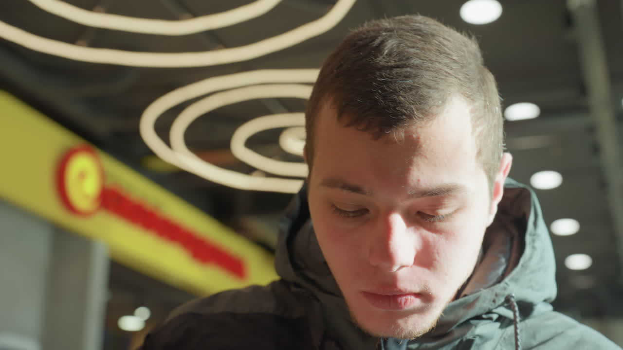 Uomo bianco che guarda in basso in una food court di un centro commerciale, indossa una giacca invernale sotto luci neon circolari, ritratto ravvicinato che cattura l'assaggio, espressioni concentrate e pensierose, azioni da studente e viaggiatore mentre lui