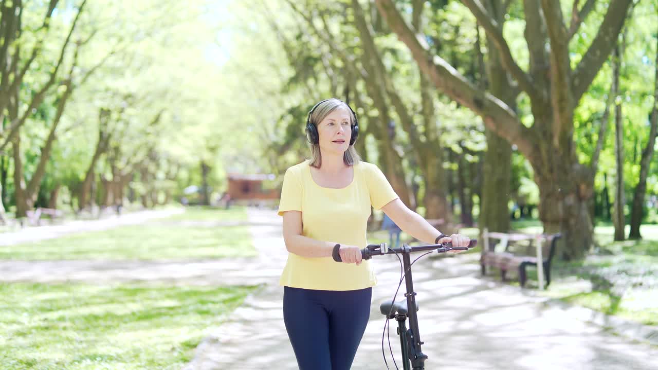 mujer activa moderna y madura feliz camina en bicicleta por el parque urbano de la ciudad con auriculares disfruta y escucha música