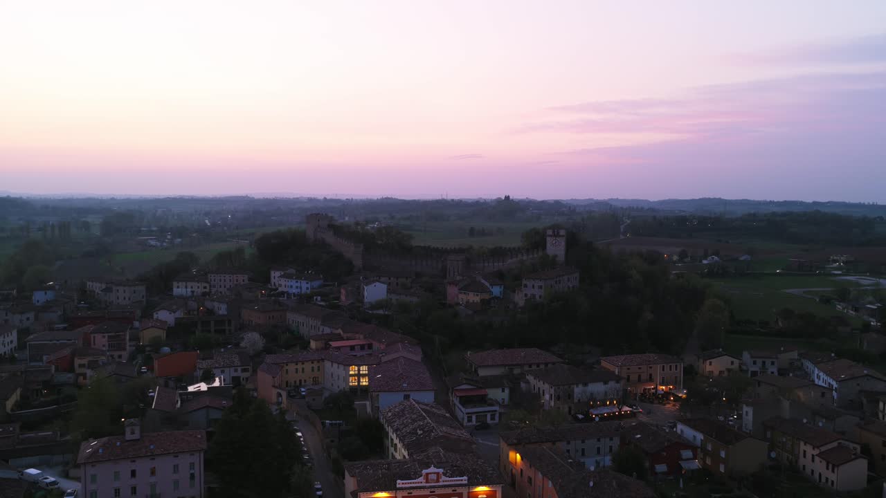Monzambano village at dusk, illuminated houses and medieval castle on hill under picturesque twilight sky, Italy. Aerial backward, copy space