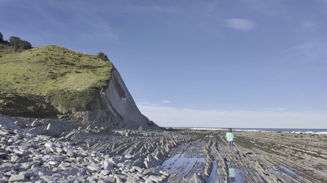 Giovane donna che esplora le incredibili formazioni di flysch sulla splendida spiaggia di Sakoneta, situata nel Geoparco della Costa Basca a Zumaia, durante una giornata di sole con cielo azzurro e limpido.
