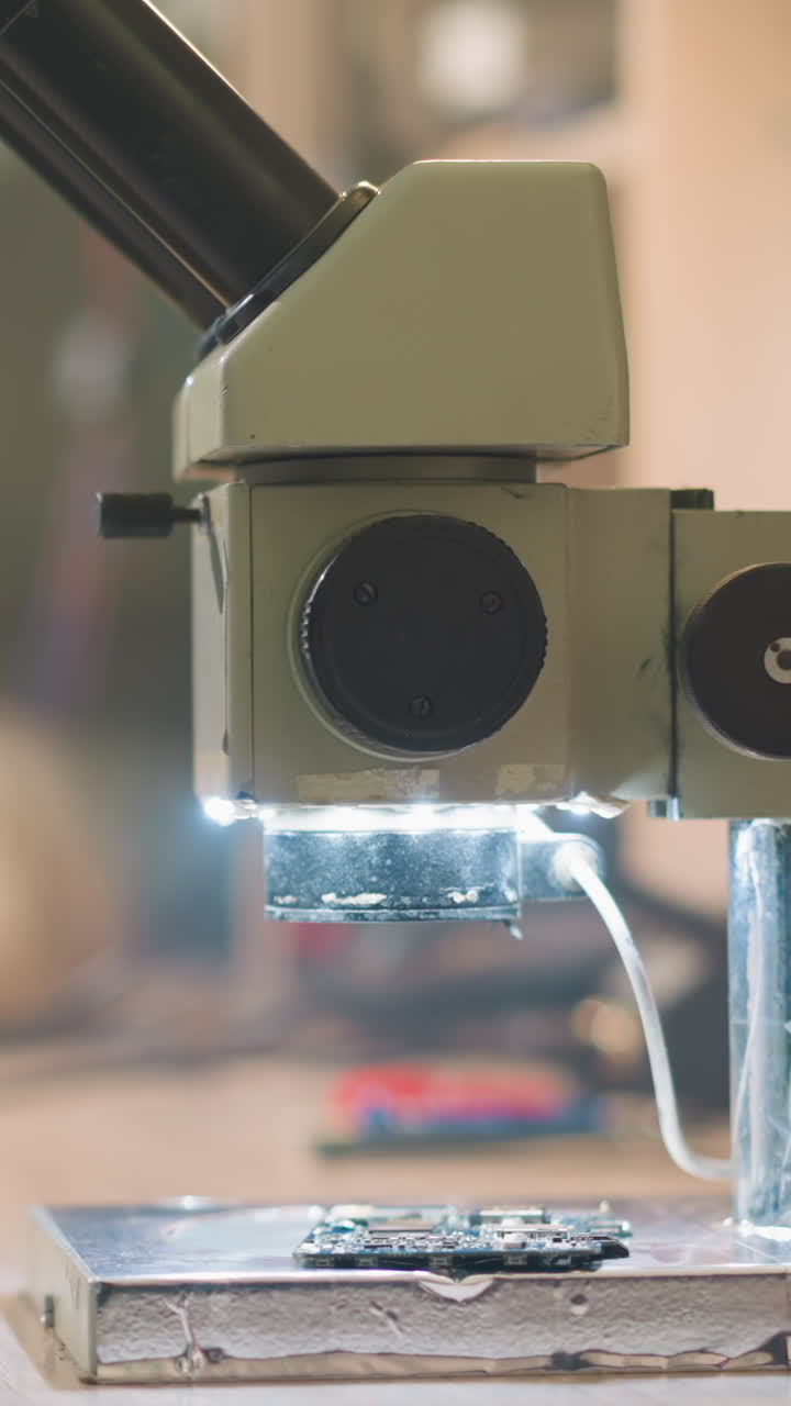 Close-up view of a microscope with LED lights illuminating a circuit board on the lab bench, a person in a lab coat and black gloves is preparing to examine the board