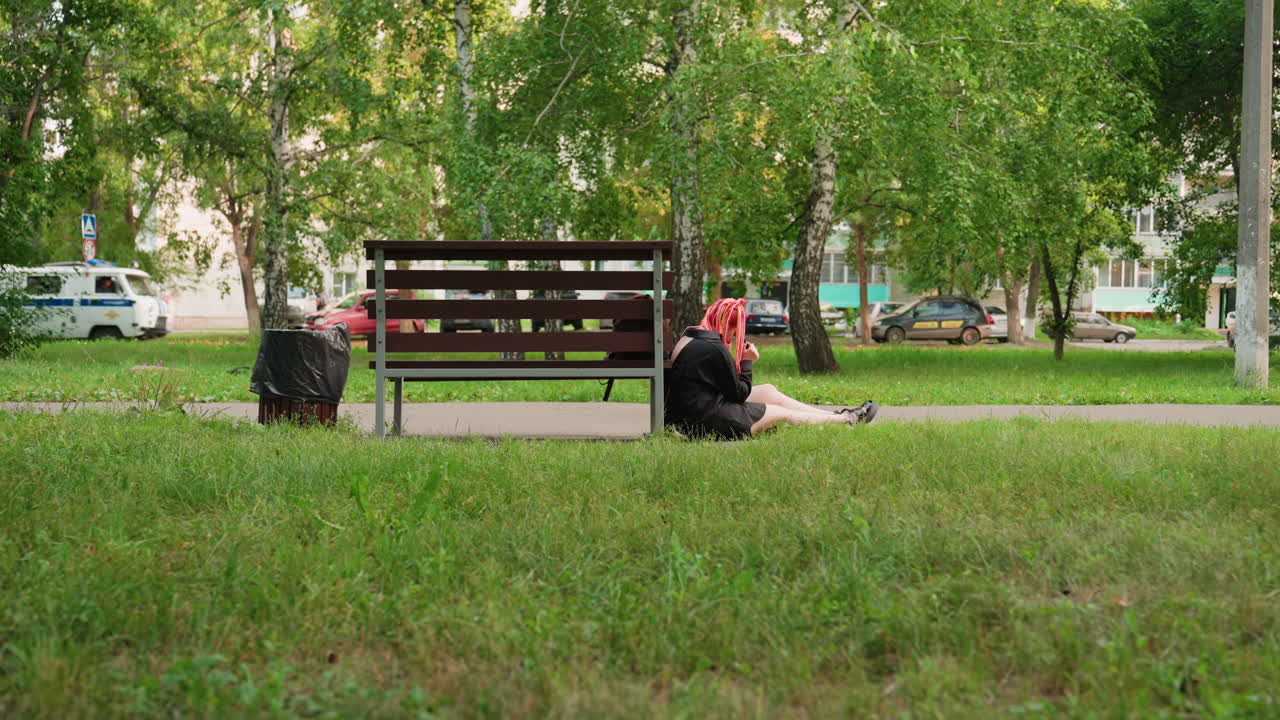 젊은 사람이 도시 공원의 나무 벤치 옆에 앉아 있고, 옆에는 백팩이 놓여 있으며, 전경에는 풀이 있고, 배경에는 자작나무가 있고, 고개를 숙여 조용히 생각에 잠겨 있으며, 우울한 분위기, 늦은 오후의 햇살, 느린 속도