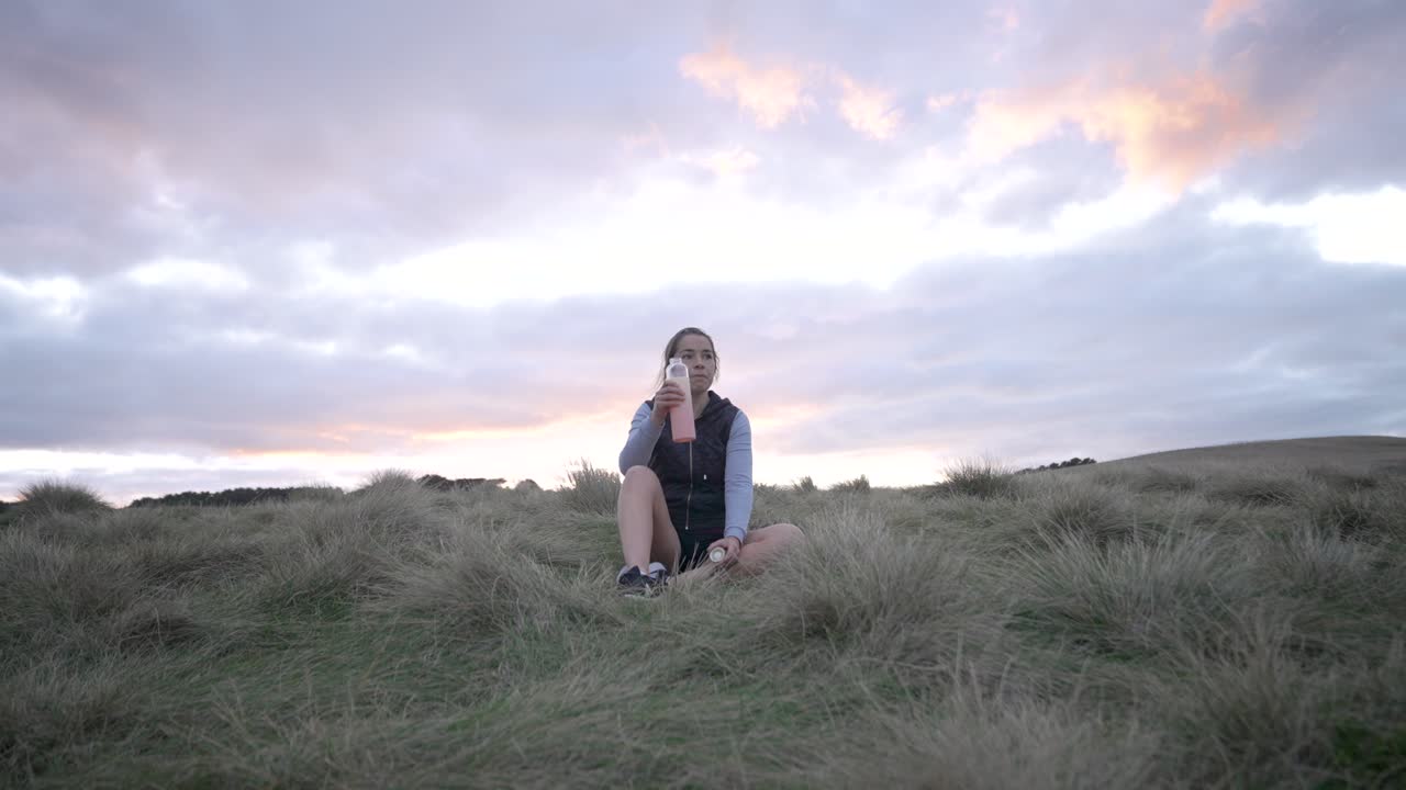 mujer joven sentada en la colina bebiendo agua después del entrenamiento amanecer detrás