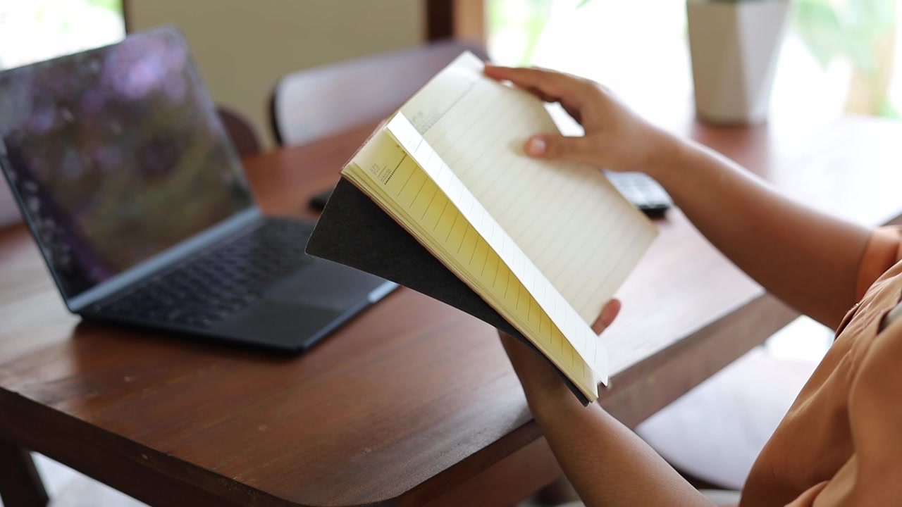An Asian woman is working remotely, sitting at a coffee shop table and focusing on reading a book, with a laptop and a cup of coffee on the table.