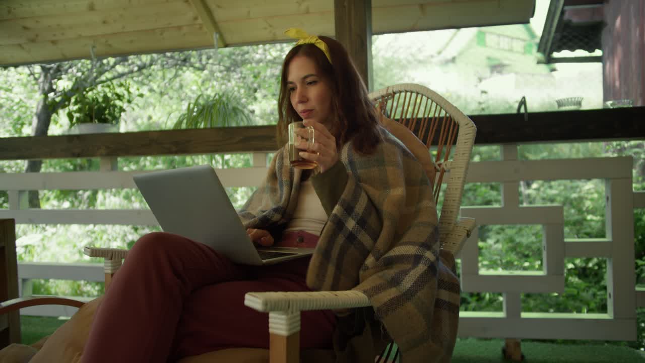 A brunette girl sits on an armchair in a plaid with tea and looks at the screen of a laptop that she has on her knees in the gazebo against the backdrop of a green forest