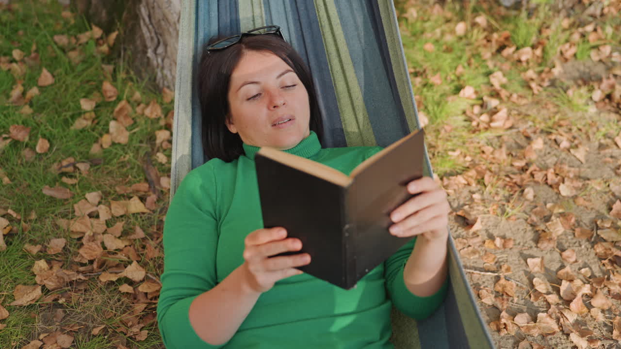 Ein abgelegener Lagerplatz bietet friedliches Lesen unter Bäumen. Eine nachdenkliche Frau liest gemächlich unter herbstlichem Blätterdach. Eine einsame Frau entspannt sich in ihrer Hängematte, während Blätter leise um sie herum fallen.