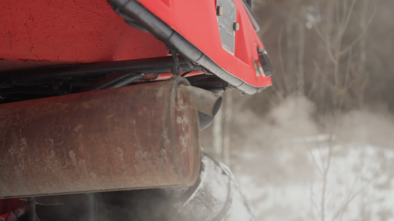 Rød ATV-udstødning frigiver hvid damp, nærbillede af rusten udstødning og udstødningsrør på frossen mark, synlig damp og vintersnavs, afdæmpet skovbaggrund, motor i tomgang, robust offroad-atmosfære.