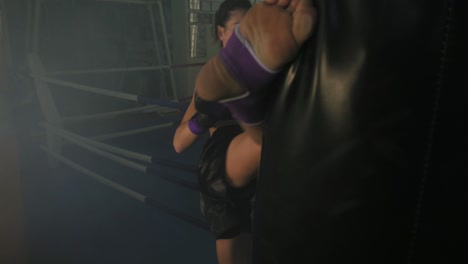 Slow-Motion-shot-of-a-leg-hit.-Strong-athletic-female-kickboxer-exercising-with-a-boxing-bag-in-dark-gym-with-smoke-and-kicking-a