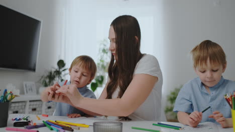 A-young-mother-with-two-children-sitting-at-a-white-table-draws-colored-pencils-on-paper-helping-to-do-homework