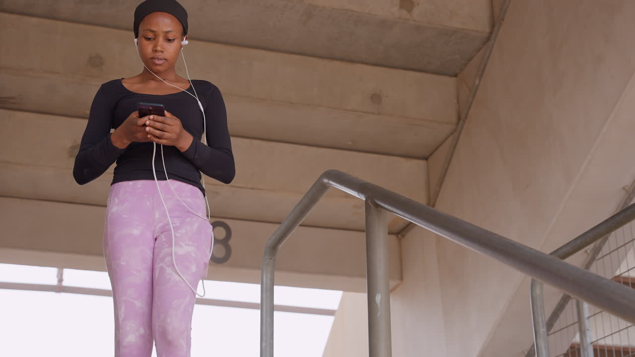 Pendolare di colore sulla scala che controlla l'orario, fermandosi sul pianerottolo con la mano sulla ringhiera, pantaloni viola casual e auricolari con filo, che dà un'occhiata al telefono per orari e aggiornamenti sui trasporti in un ambiente di cemento.