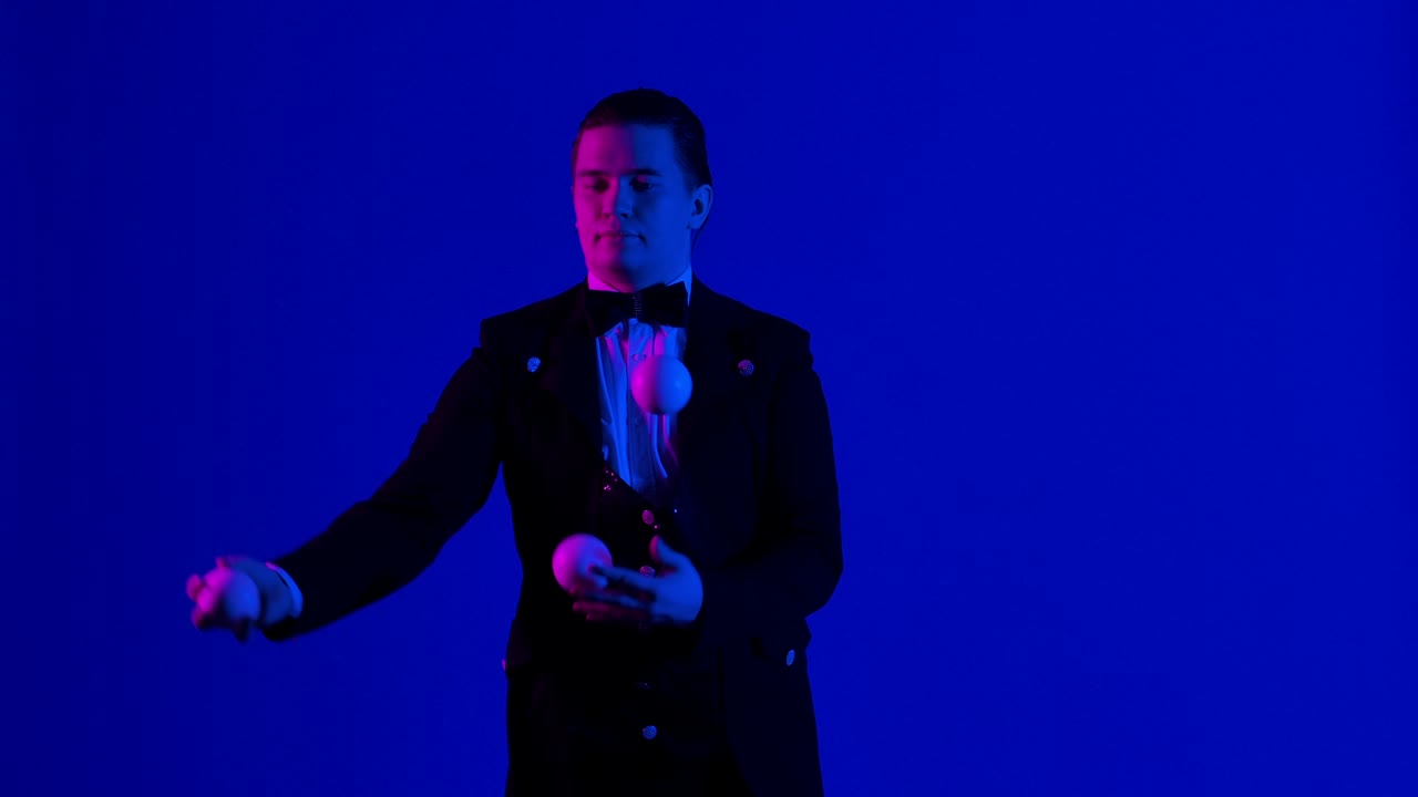 Man juggler shows tricks with white balls, tossing and catching them. Stylish showman in black suit practices concentration, dexterity and skill. Blue screen, chroma key. Close up. Slow motion