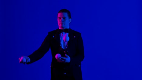 man juggler shows tricks with white balls, tossing and catching them. stylish showman in black suit practices concentration, dexterity and skill. blue screen, chroma key. close up. slow motion