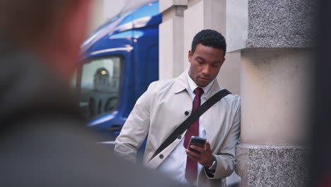 Joven-Hombre-De-Negocios-Negro-Apoyado-En-Una-Pared-En-La-Calle-De-Londres-Usando-Su-Teléfono-Inteligente,-Enfoque-Selectivo,-Cintura-Para-Arriba