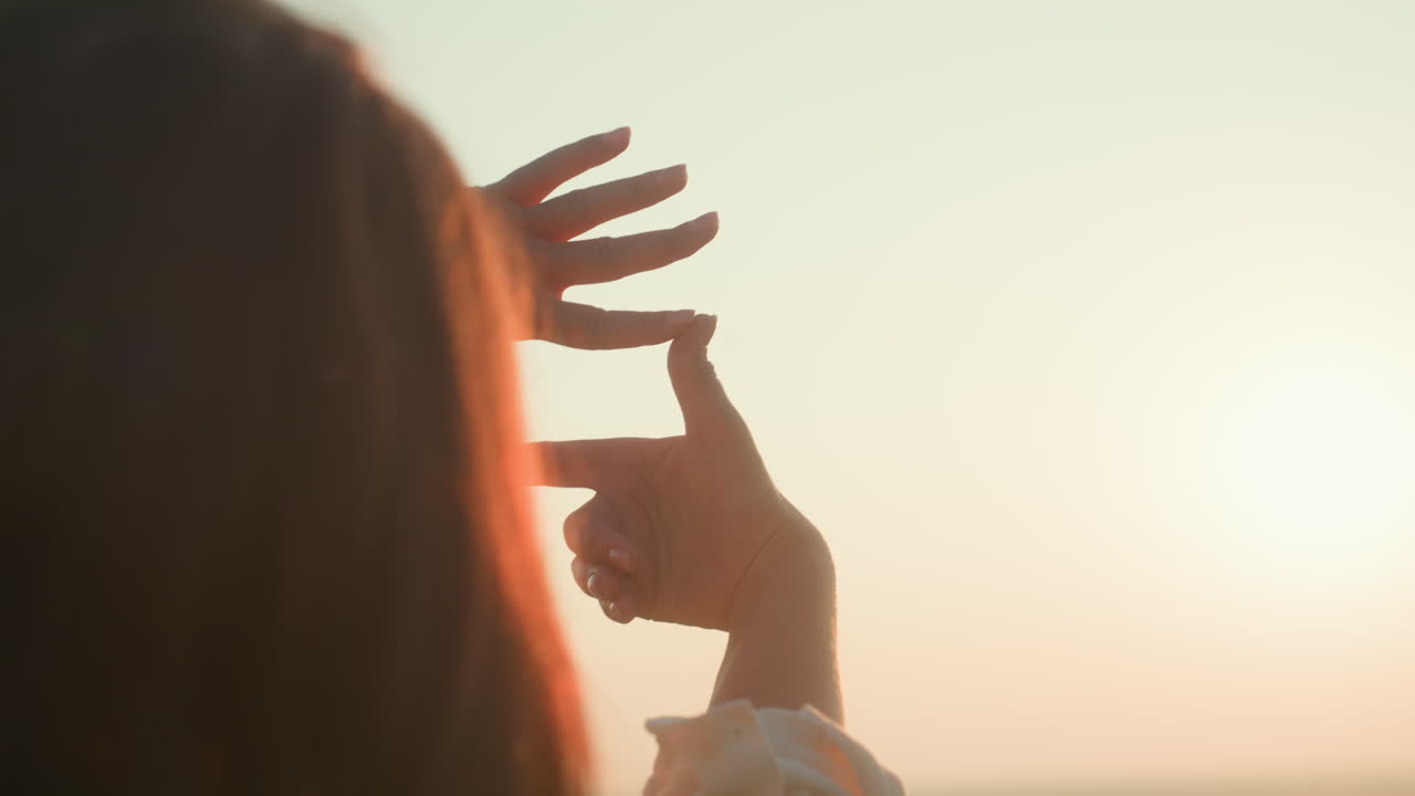 Rear view of woman making subtle hand frame gesture over glowing sunset river and wild field, warm golden backlight highlighting long hair and fingers