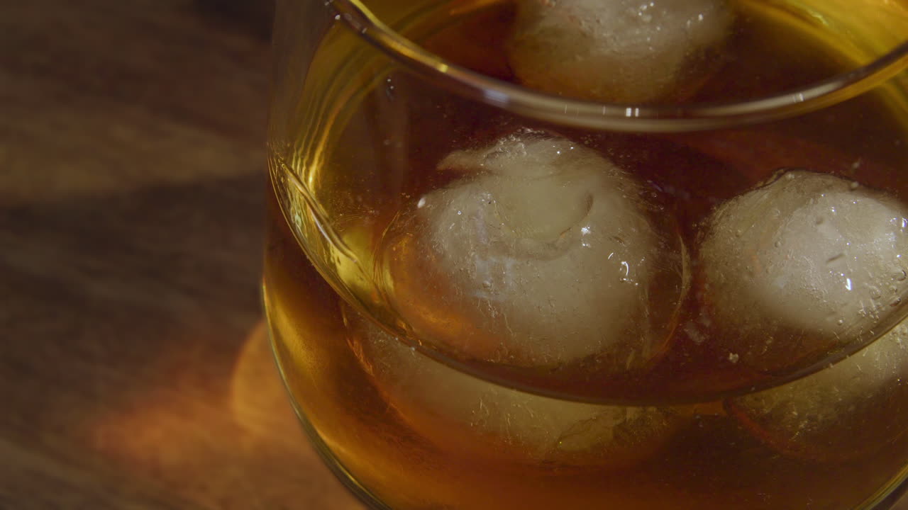 A male hand places a glass with whiskey on a wooden surface with two ice cubes inside