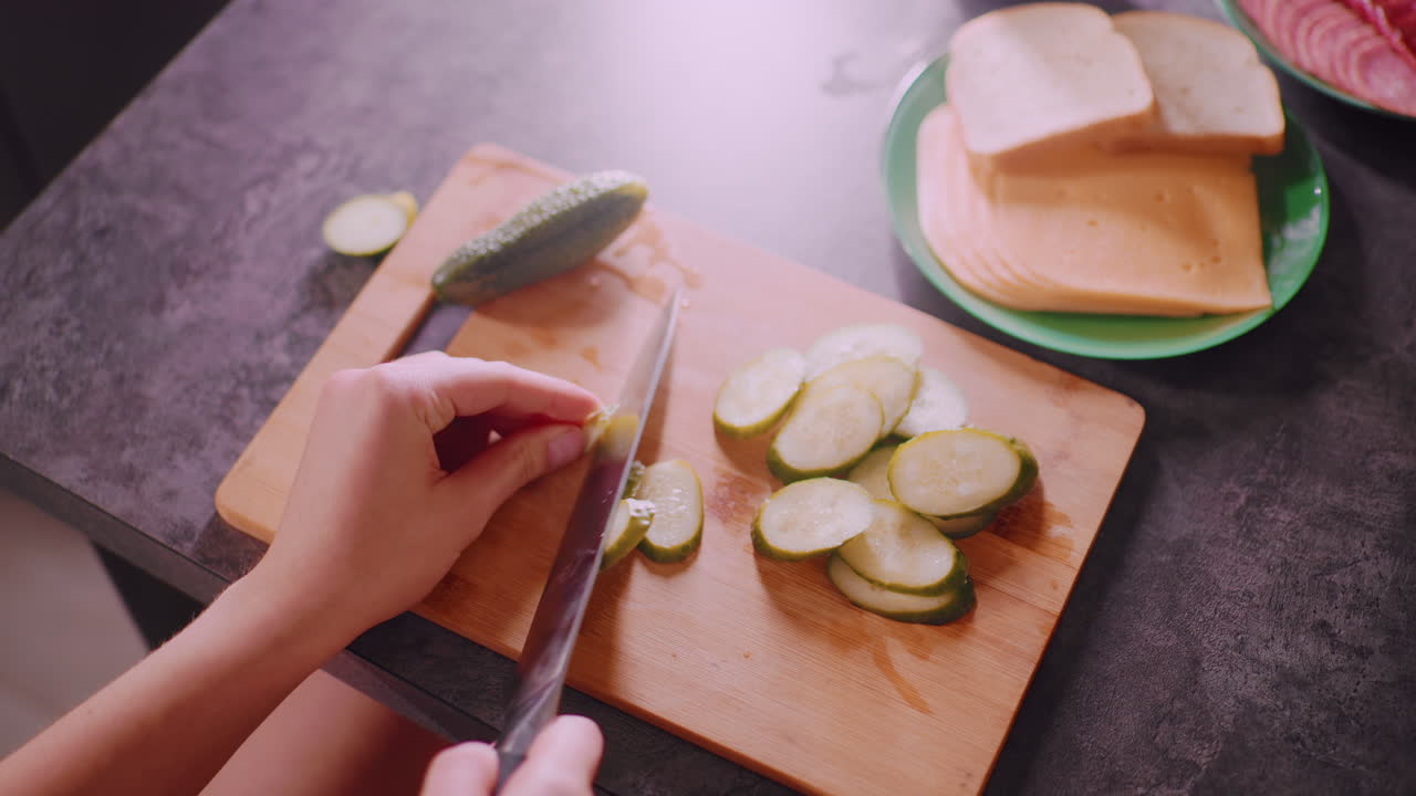 Overhead view of chef slicing cucumber on wooden cutting board beside green plate with bread and cheese slices, salami arranged nearby, and coffee cup on countertop under warm sunlight