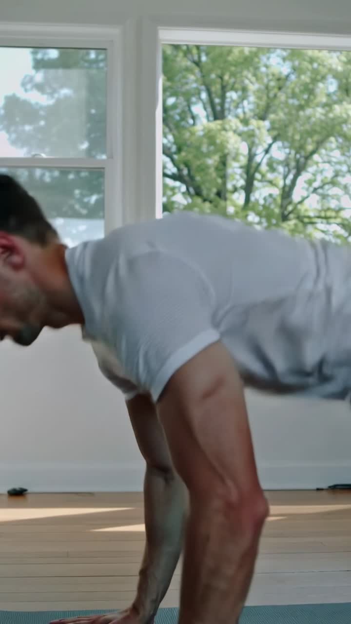 Video verticale: Ragazzo con maglietta grigia che inizia uno squat e fa plank push-up, con finestre di casa, pavimento e telefono, spazio per il testo.